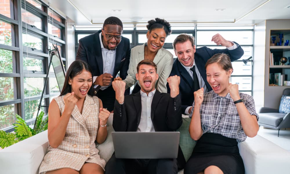 Six people crowd around an open laptop computer, delighted with what they are seeing